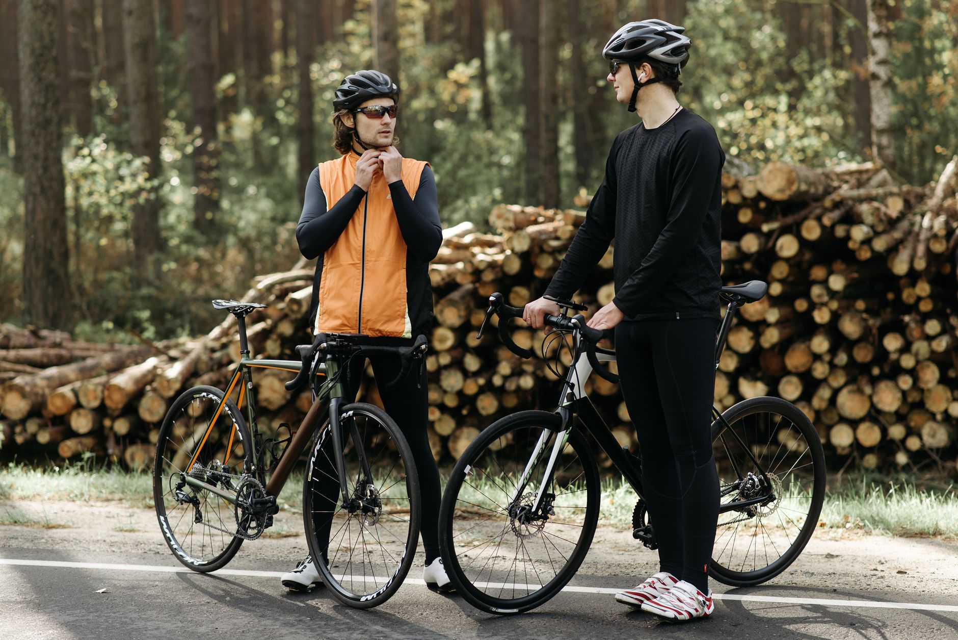 Two male cyclists pause on a forest road beside stacked logs, wearing helmets and athletic gear.