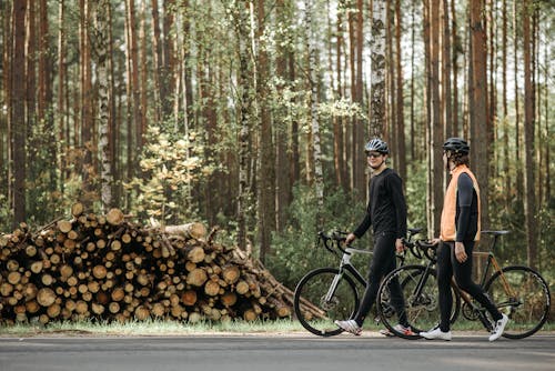 Men Walking While Holding Their Bicycle Near Wood Logs 