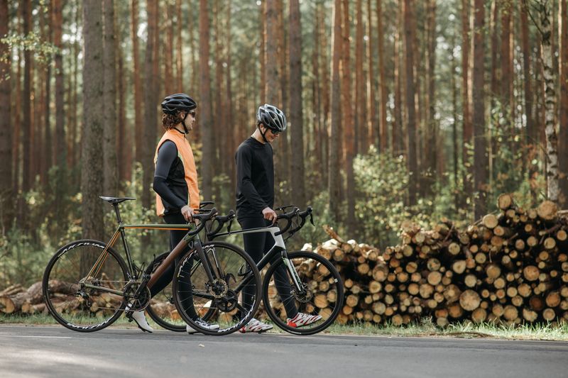 Two cyclists in helmets walking their road bikes alongside a wooded area.
