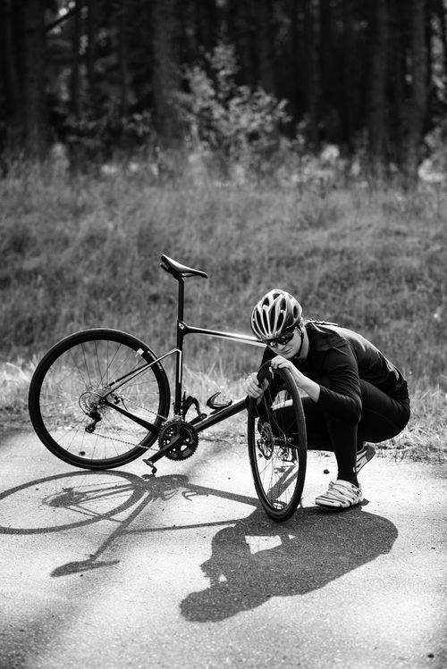 Free A cyclist in gear repairs their bike on a sunlit outdoor path, captured in black and white. Stock Photo