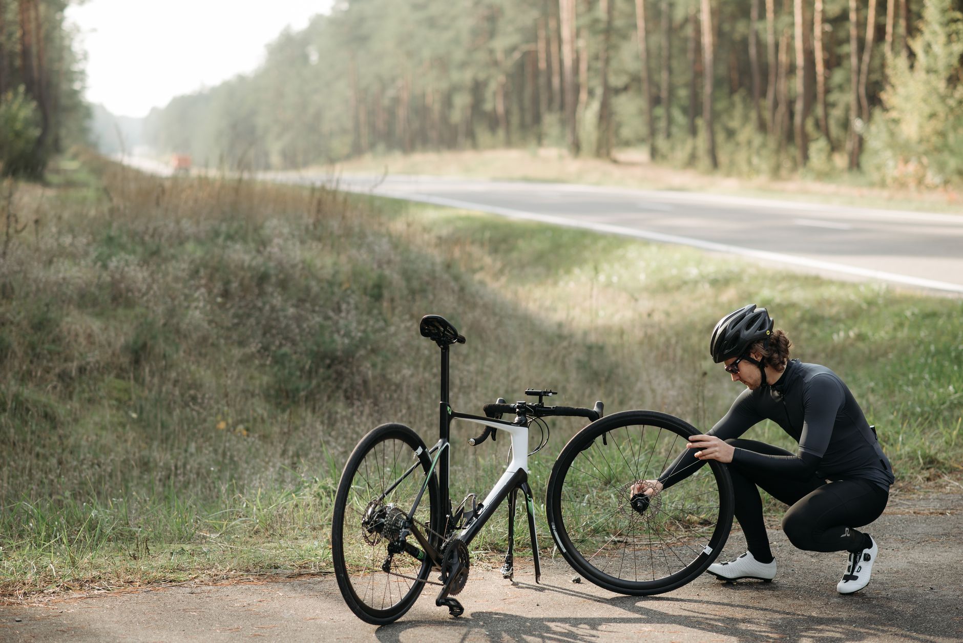 Cyclist fixing a bicycle wheel on a rural roadside surrounded by forest.