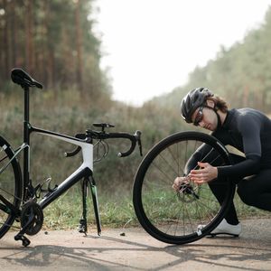Cyclist in black kit crouched by a quiet forest road, working on the back wheel of a road bike