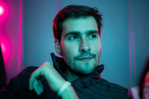 Confident brutal male with brown eyes sitting in room with colorful neon light and looking away