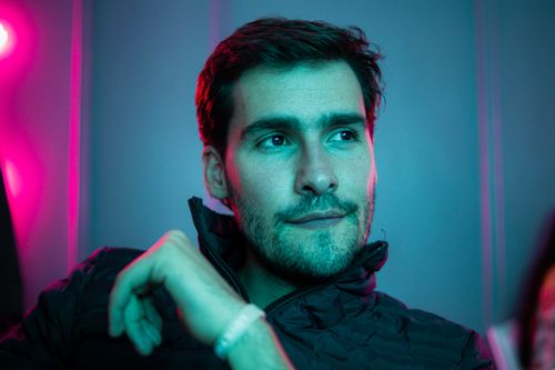 Free Confident brutal male with brown eyes sitting in room with colorful neon light and looking away Stock Photo