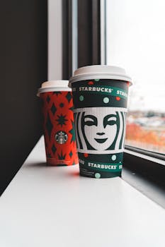 Two Starbucks holiday-themed coffee cups on a bright window sill.
