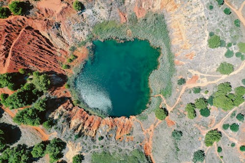 Drone view of turquoise water of small pond surrounded by rough stony formation with green plants