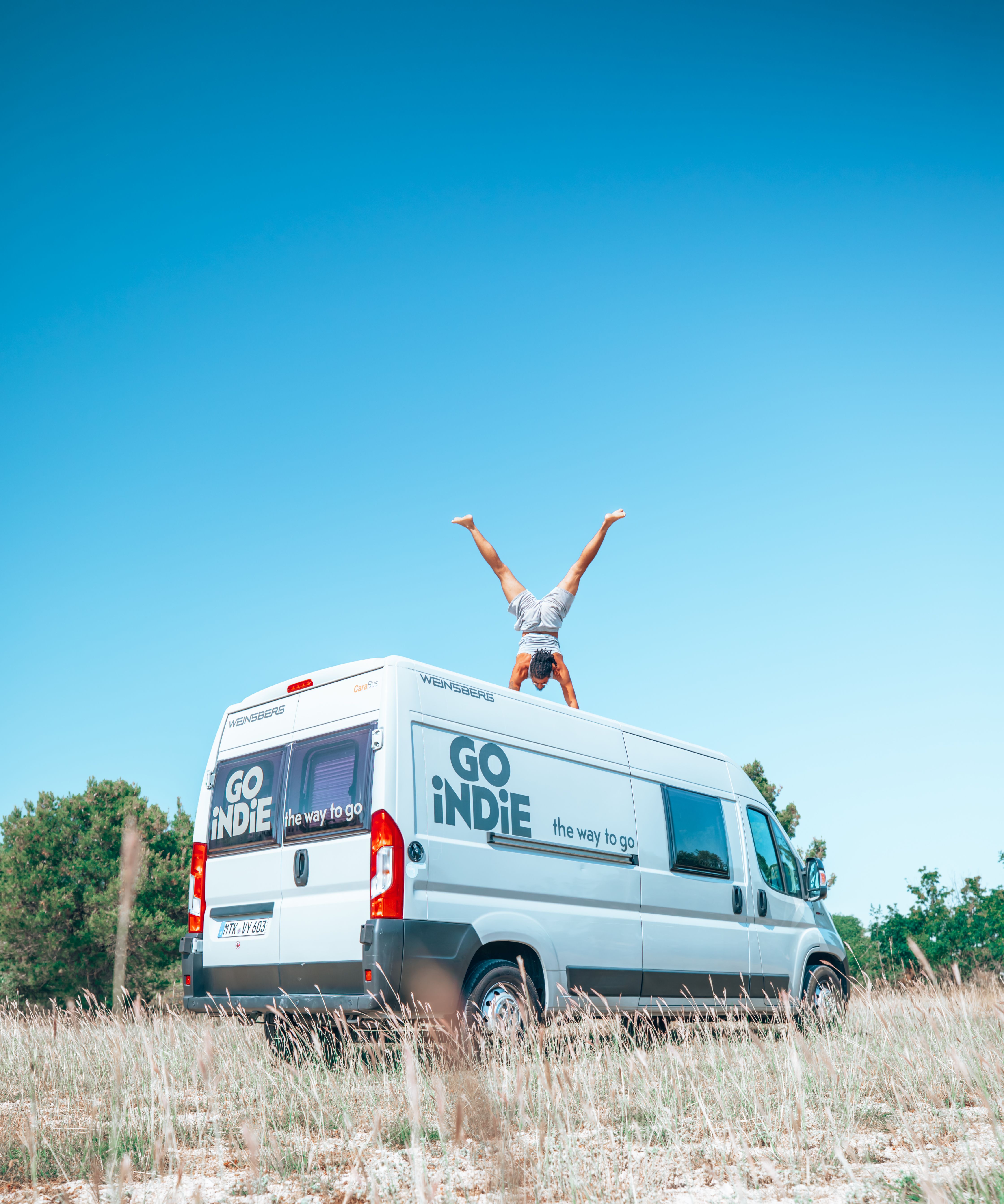 A person performs a handstand on a camper van roof under a clear blue sky outdoors.