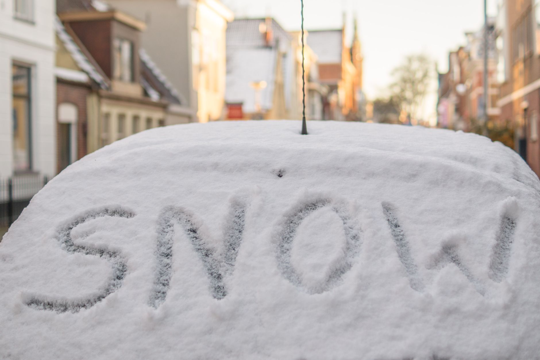 A snow-covered car with the word 'SNOW' written on it, parked on a winter street lined with buildings.