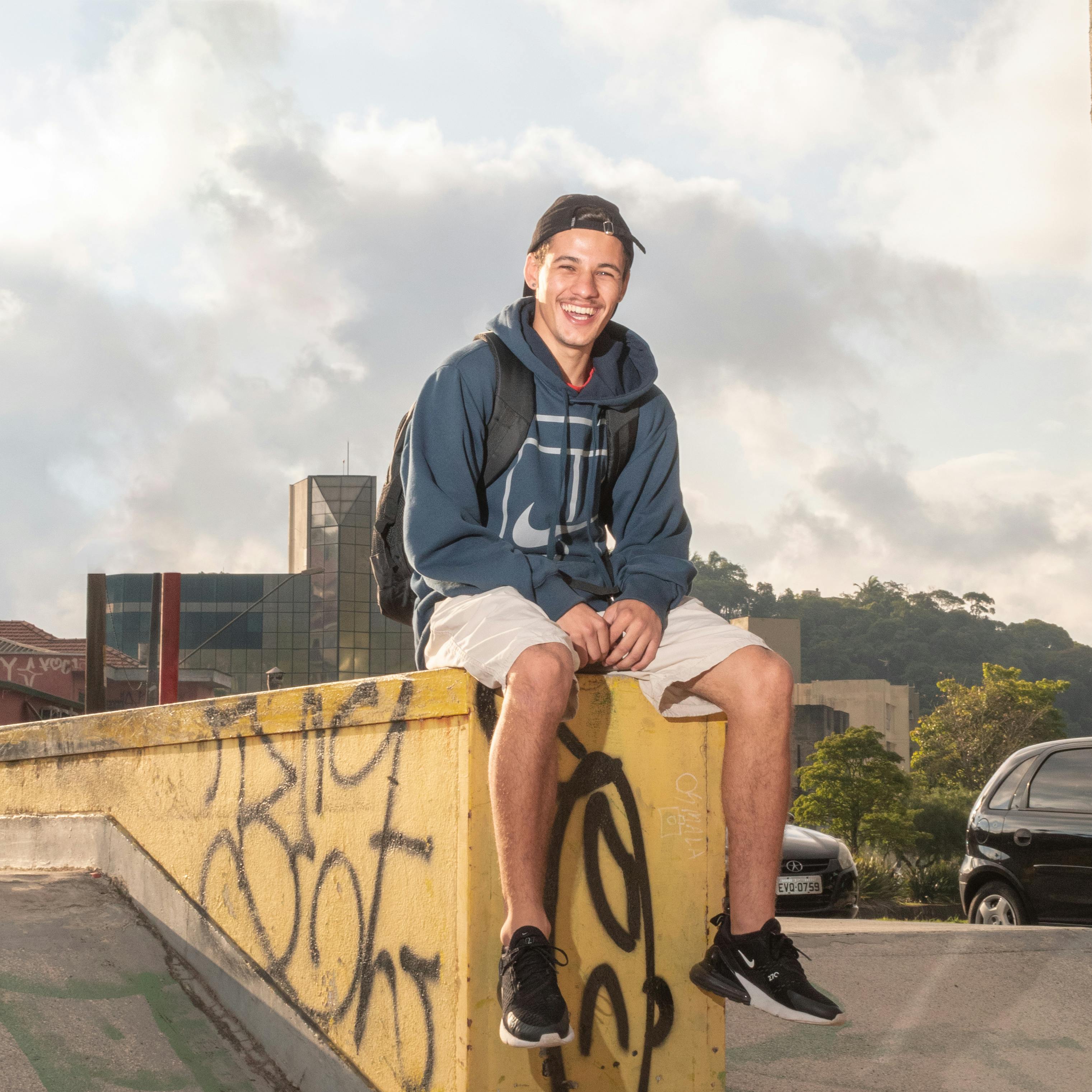 Casually dressed young man with a backpack sitting on a graffiti-covered wall outdoors.