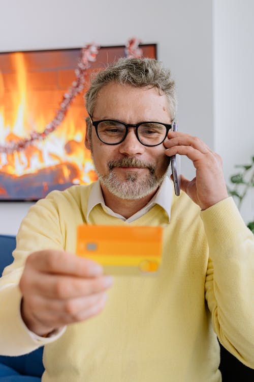 Man in Yellow Sweater Wearing Black Framed Eyeglasses Holding a Card While Talking on Phone