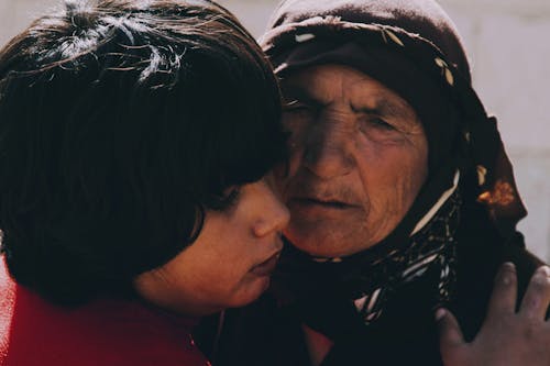 Crop ethnic child embracing unhappy elderly grandma in headscarf while looking away in sunlight