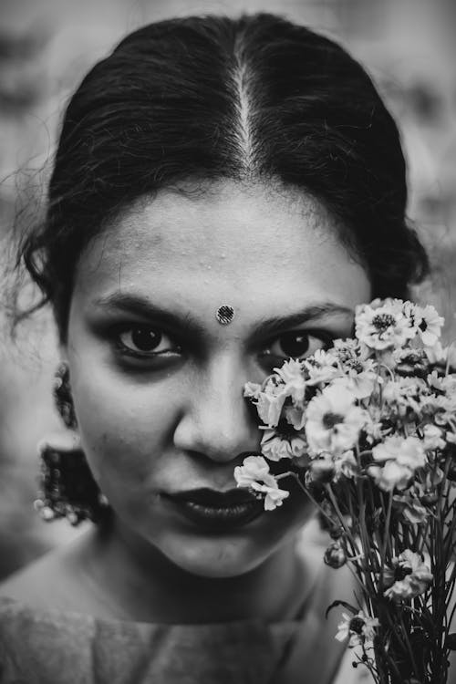 Black and white of confident young Indian female with dark hair and traditional bindi on forehead looking at camera with bunch of fresh flowers in hand