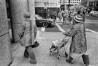 Black and white of faceless passersby in similar clothes walking on city street near road with retro cars