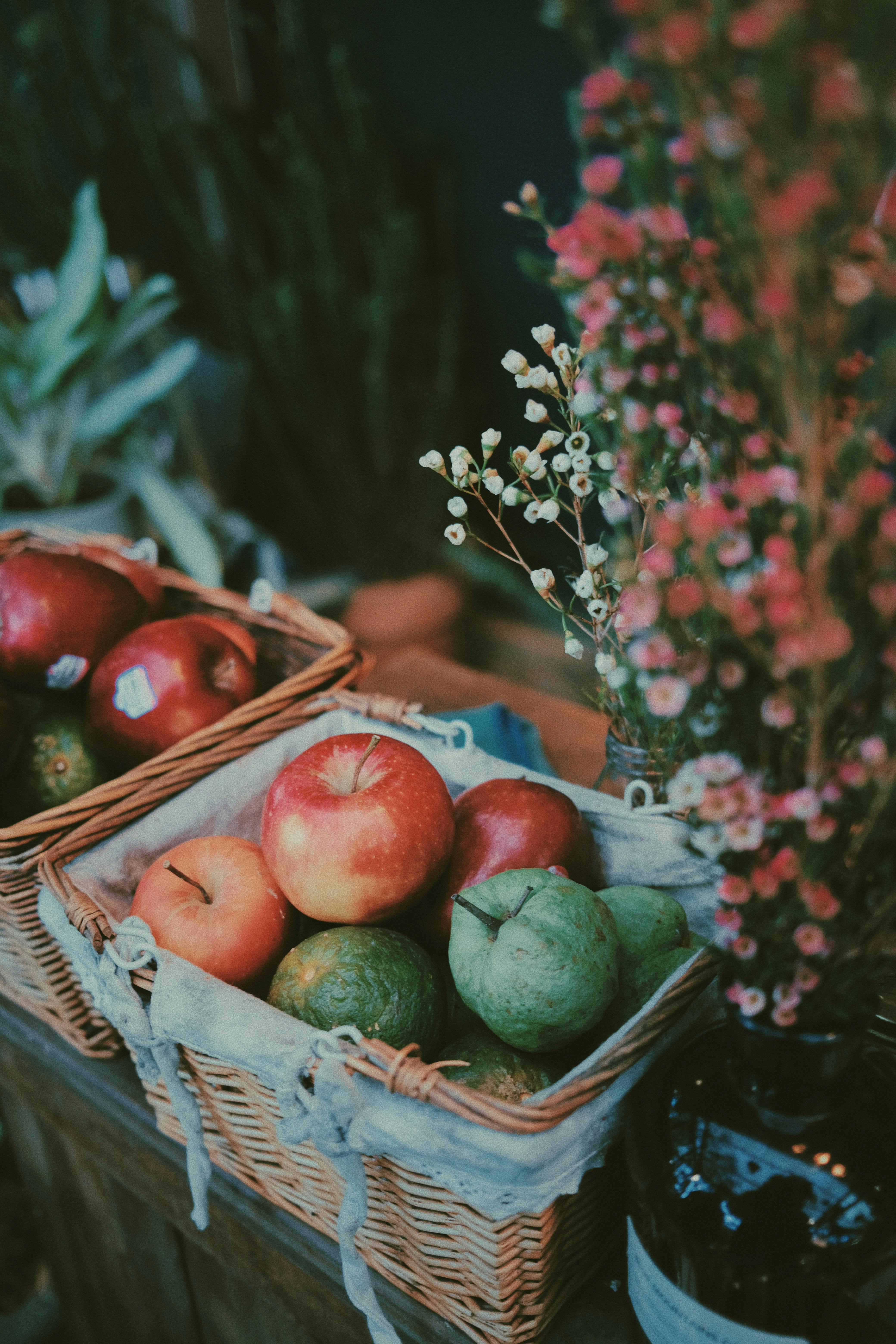 Wicker baskets filled with fresh colorful fruits placed near flowers in vase on market in daylight