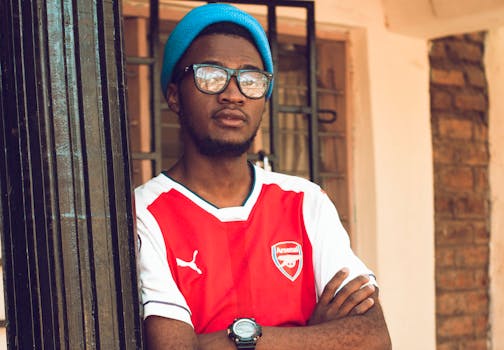 Confident man wearing Arsenal jersey and beanie, leaning against a wall with crossed arms.