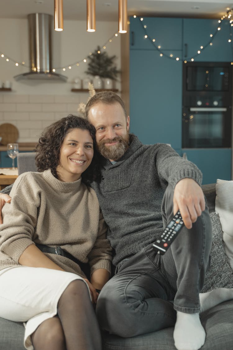 A Couple Wearing Sweaters And Sitting Side By Side Watching TV