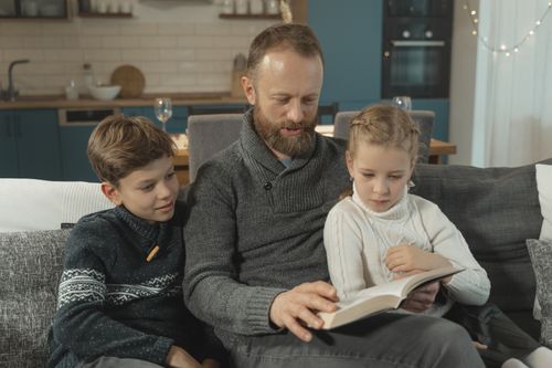 Free A father reading a book with his two children on a sofa in a warm indoor setting. Stock Photo