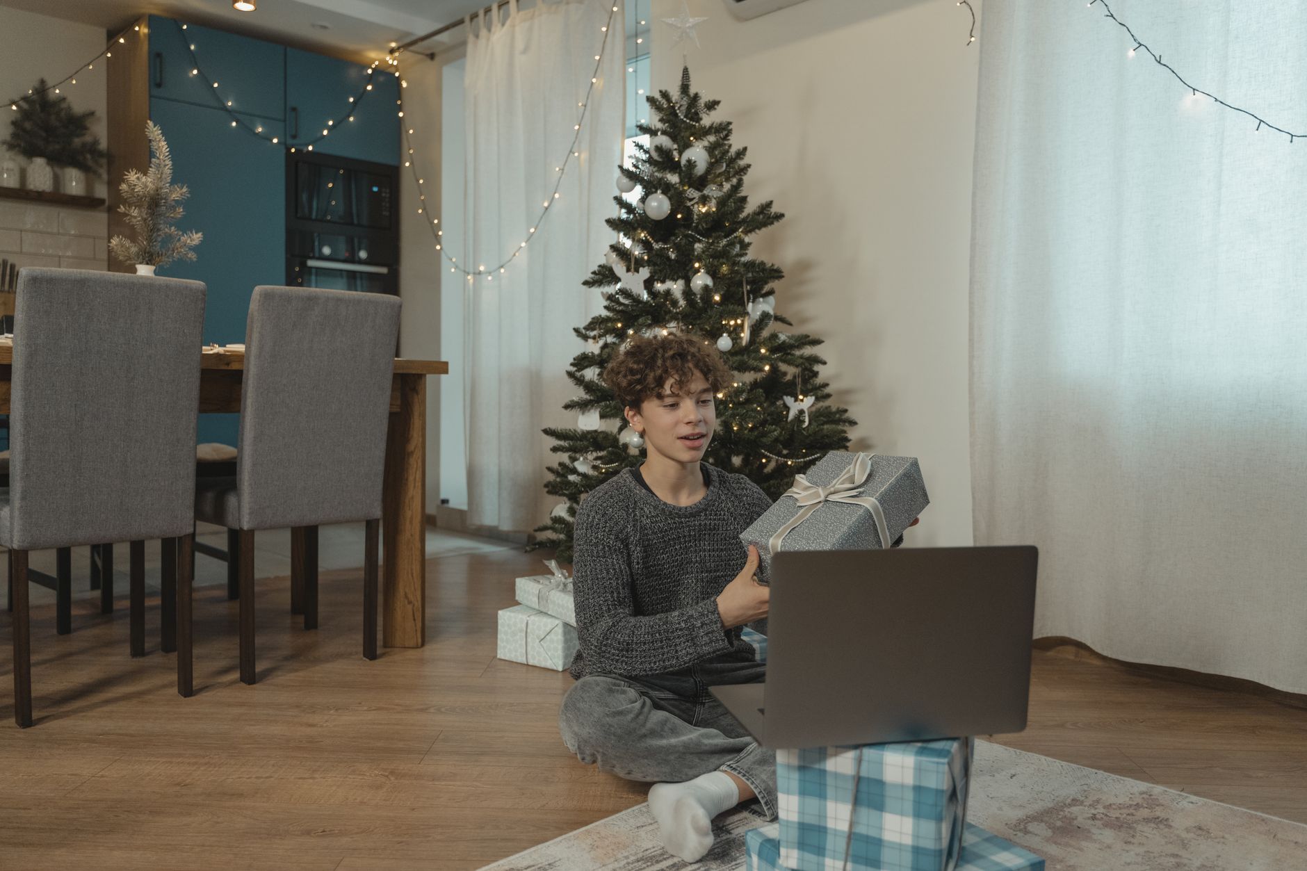 Teen boy participates in a virtual Christmas gift exchange from home, seated by a decorated tree holding a present.