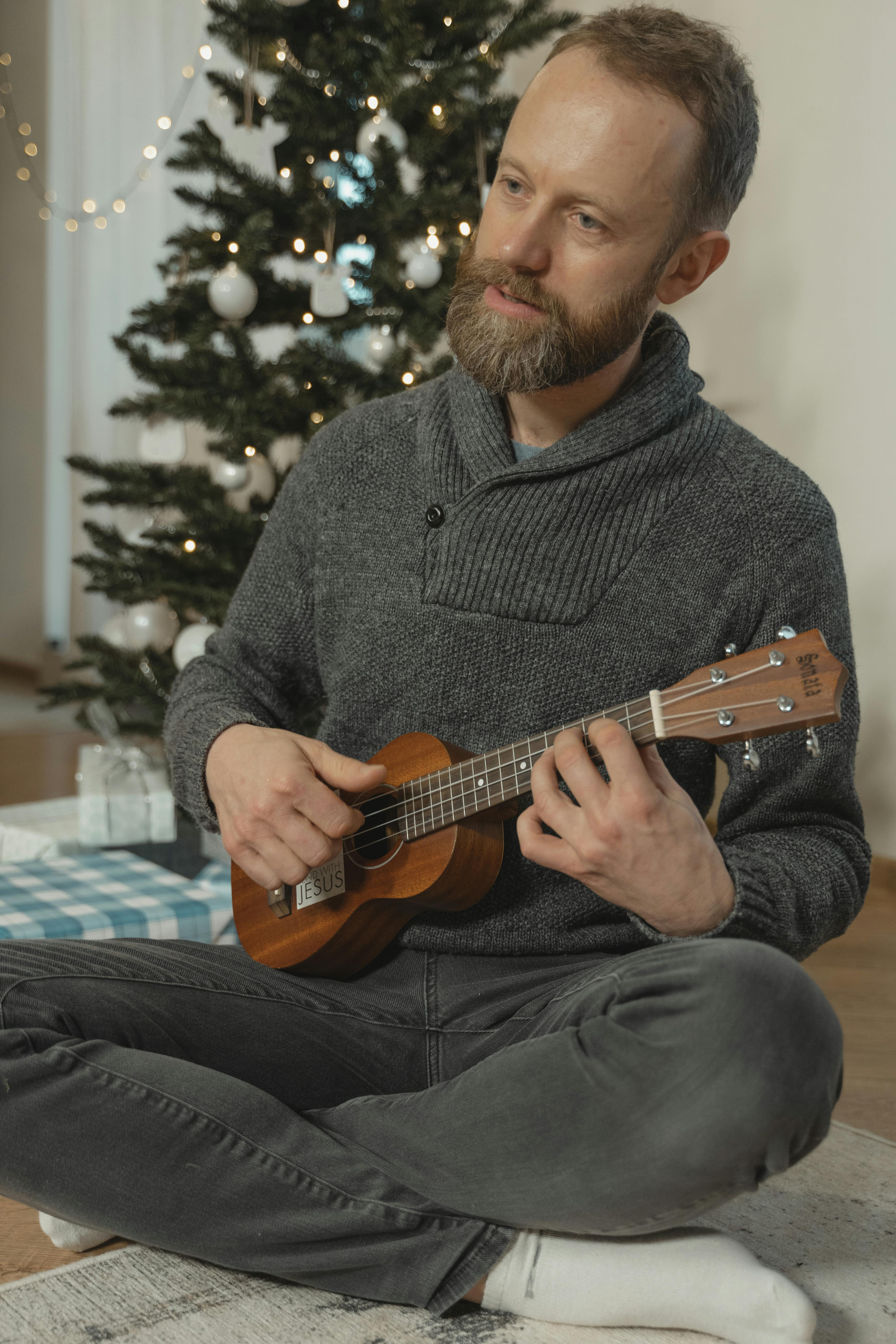 A man sits playing a ukulele next to a decorated Christmas tree at home.