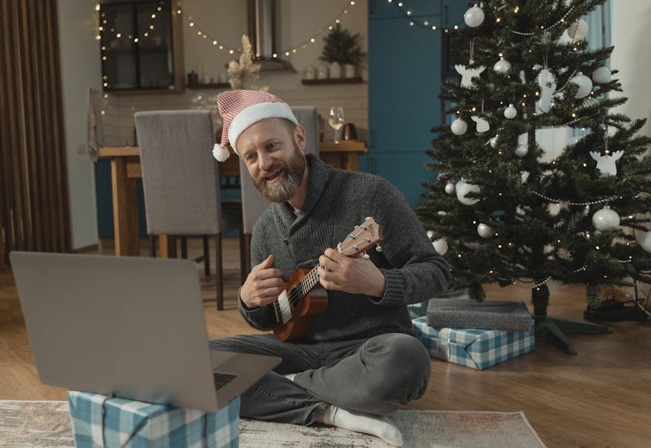 A man wearing a Santa hat plays a ukulele during a Christmas video call, sitting near a decorated tree.