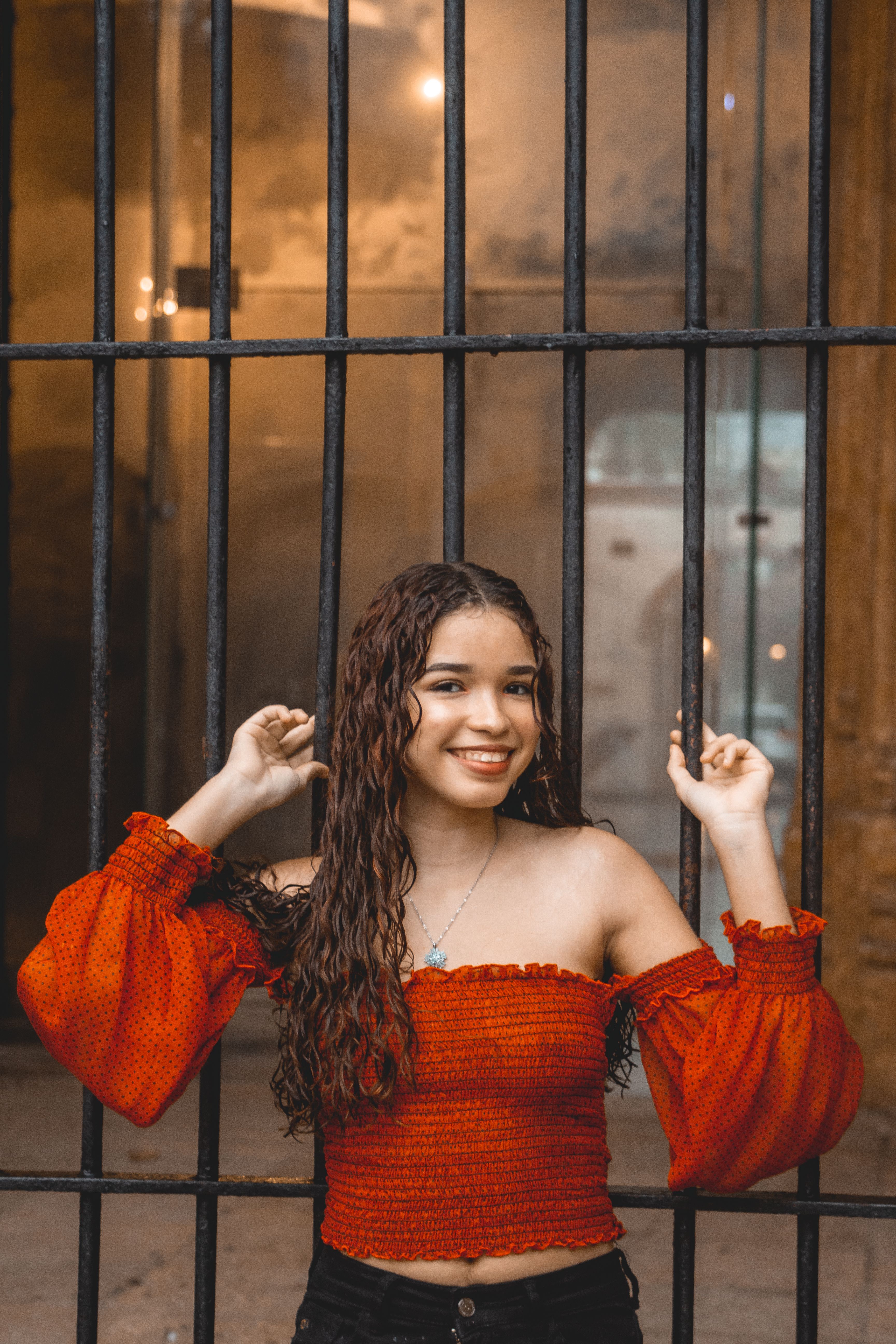 A smiling young woman with curly hair in a red off-shoulder top poses at a metal gate in Santo Domingo.