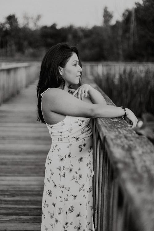 Grayscale Photo of Woman in Floral Dress Standing on Wooden Bridge 