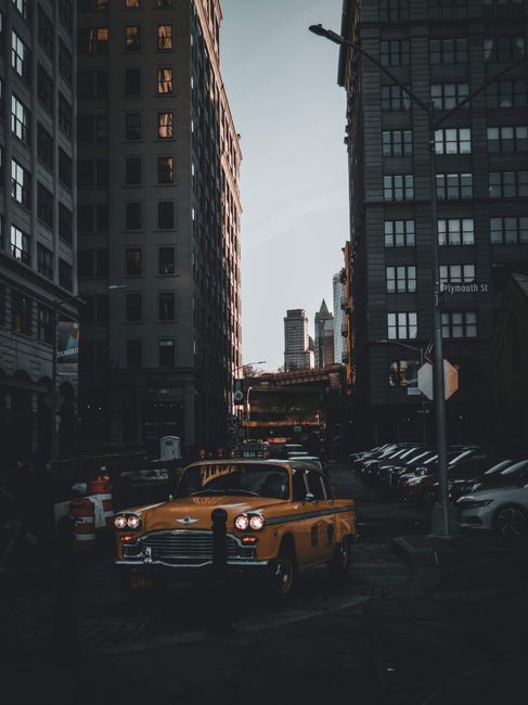 A vintage yellow cab drives through a bustling downtown New York City street framed by high-rise buildings.
