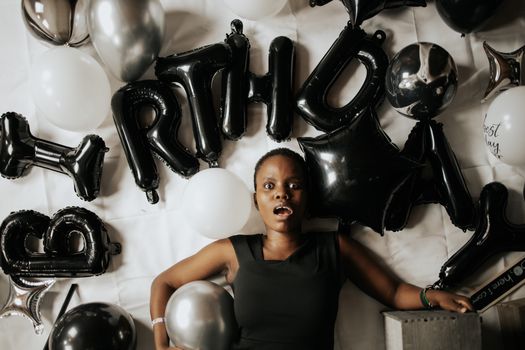 A surprised black woman surrounded by black and white balloons during her birthday celebration.