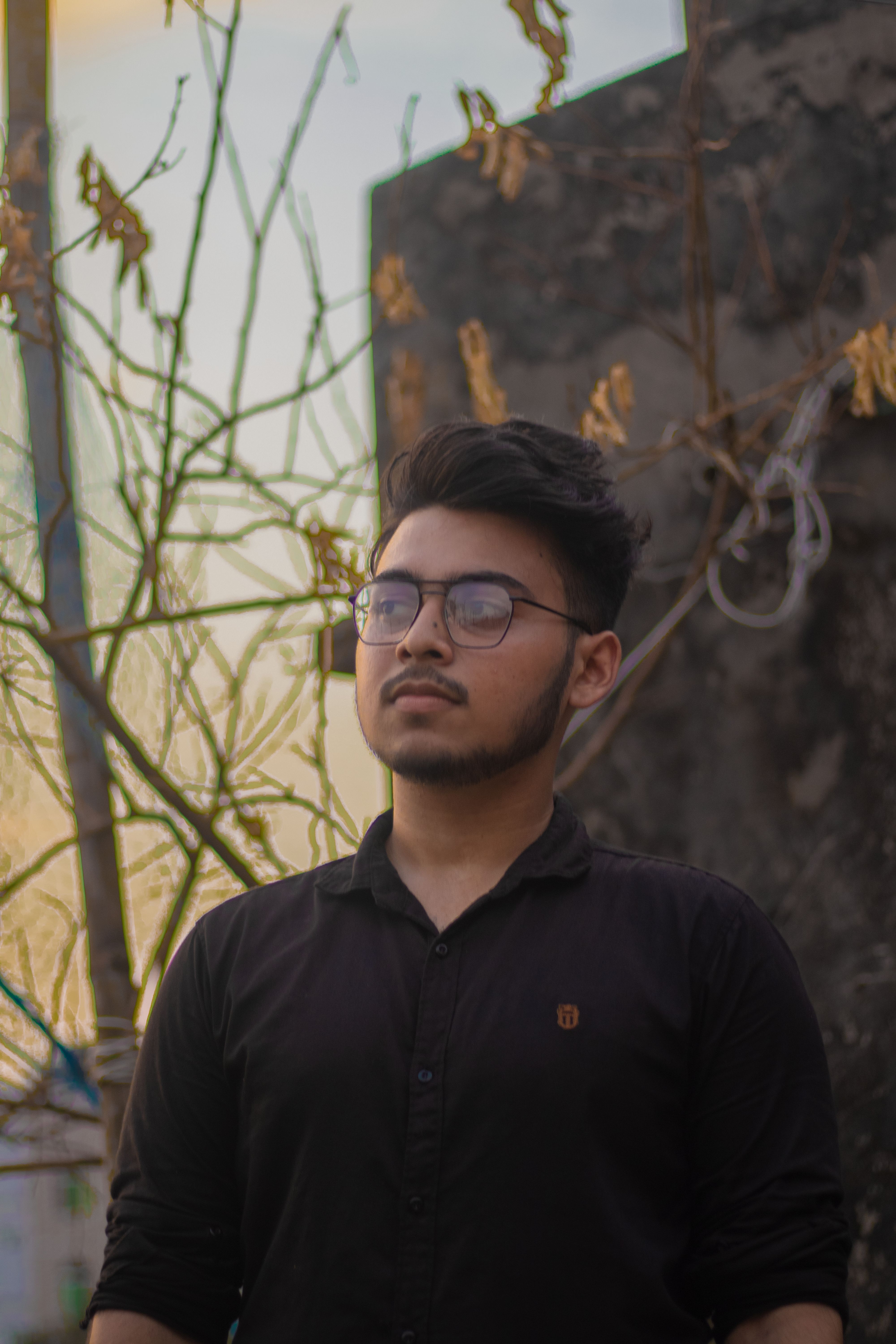 Bearded man in black, wearing glasses, gazing with intent outdoors. Bangladesh setting.