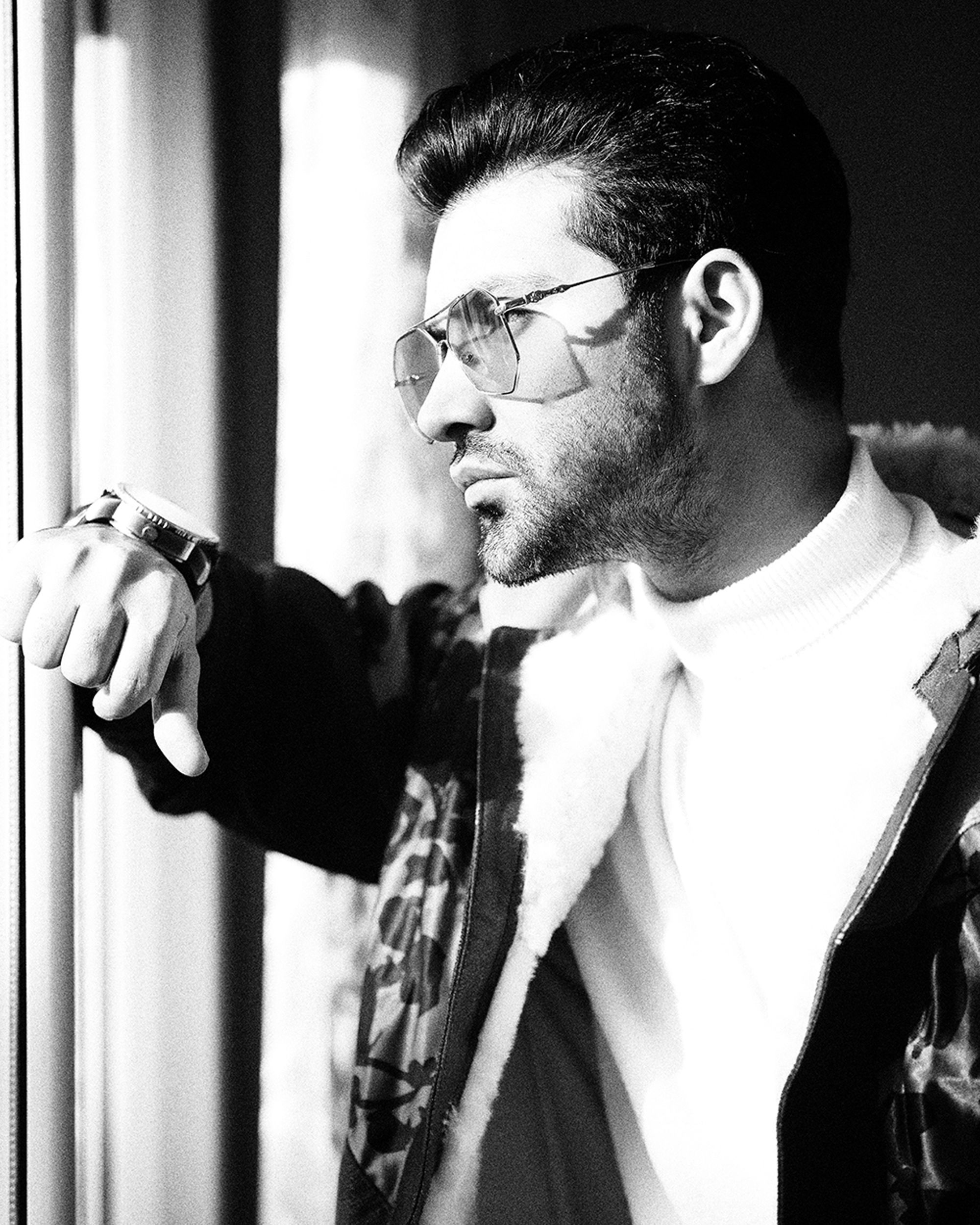 A fashionable man with eyewear and a beard posed seriously by a window in Tehran.