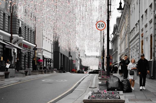 A bustling Moscow street adorned with festive hanging lights, featuring pedestrians and a 20 km/h sign.