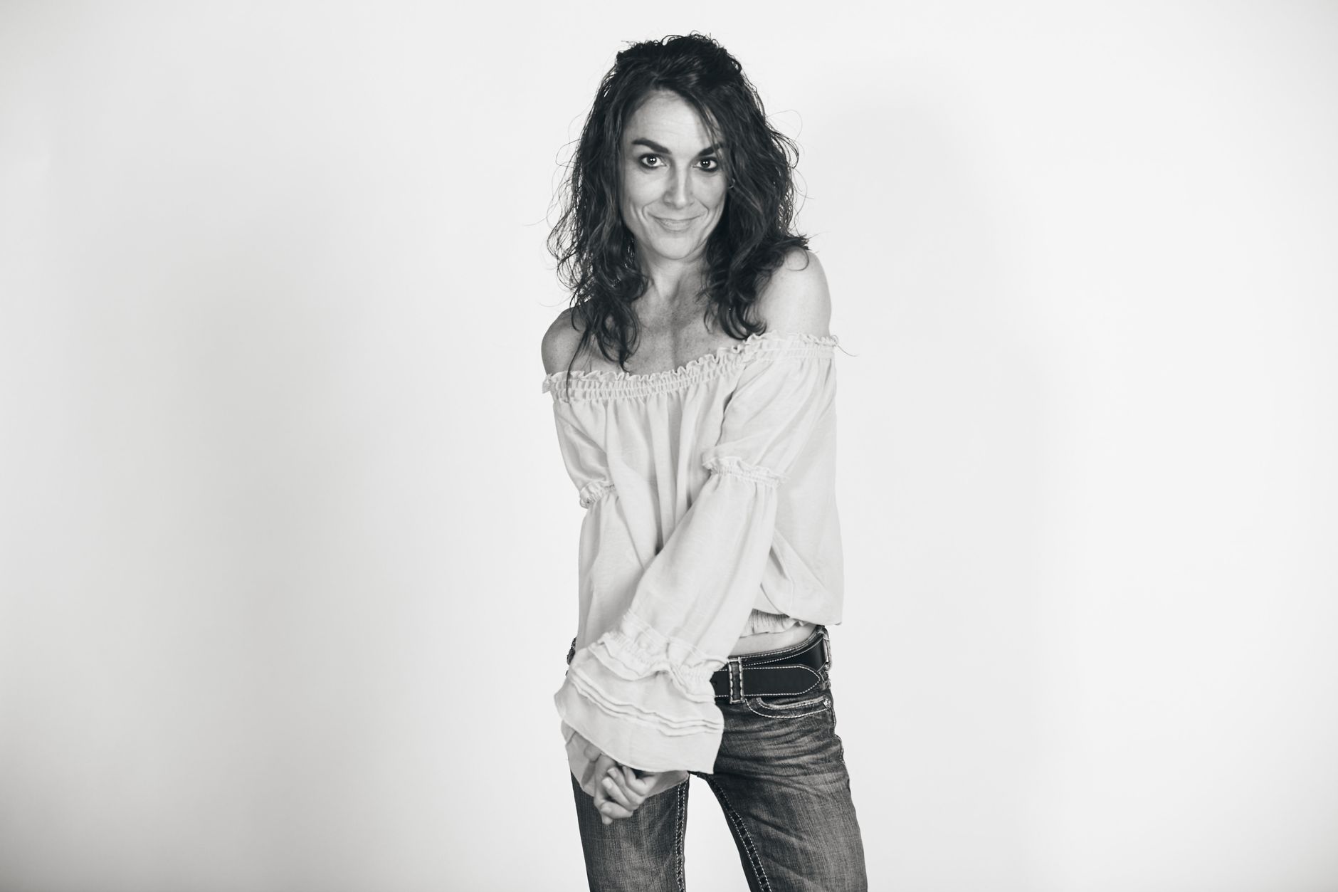 Black and white portrait of a smiling woman in casual off-shoulder top and jeans, captured in a studio setting.