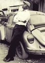 Grayscale Photo of Man in Long Sleeves and Pants Wearing Hat Leaning on Car