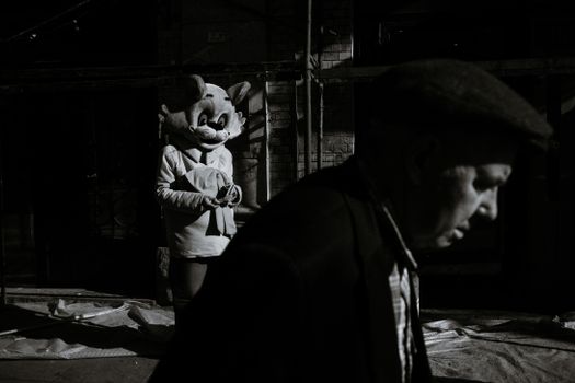 Black and white side view of elderly man in casual clothes walking near actor in costume of cat standing on polyethylene against old brick building with scaffolding in megapolis