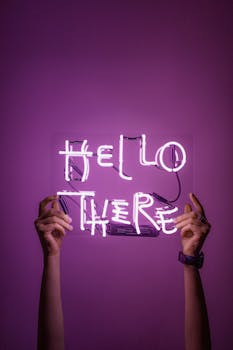Bright neon sign saying 'Hello There' held by hands against a purple background.