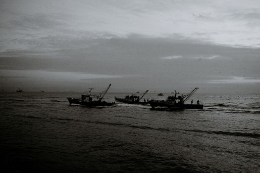 Black and white of unrecognizable fishermen on trawling boats with fishing nets on rippled ocean under cloudy sky