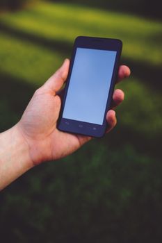 Close-up of a hand holding a blank smartphone outdoors with green blurred background.
