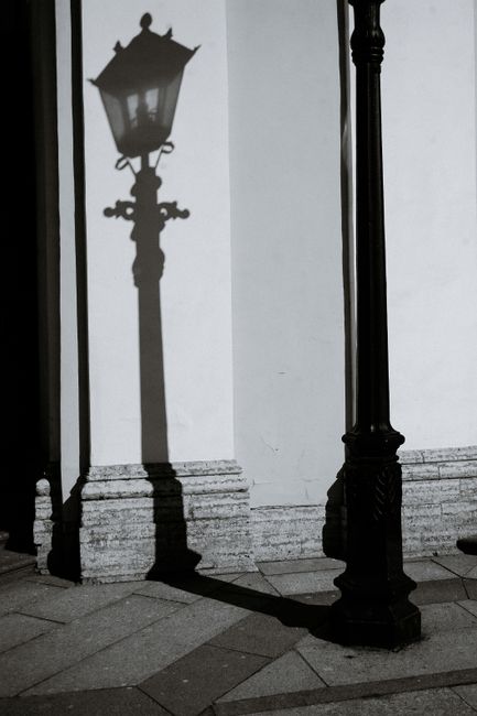Black and white of ornamental lamp shade on house wall near urban tiled pavement in sunlight
