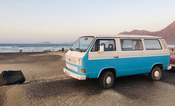 A blue vintage Volkswagen van parked on a sandy beach in Teguise, Spain, with a scenic ocean backdrop.