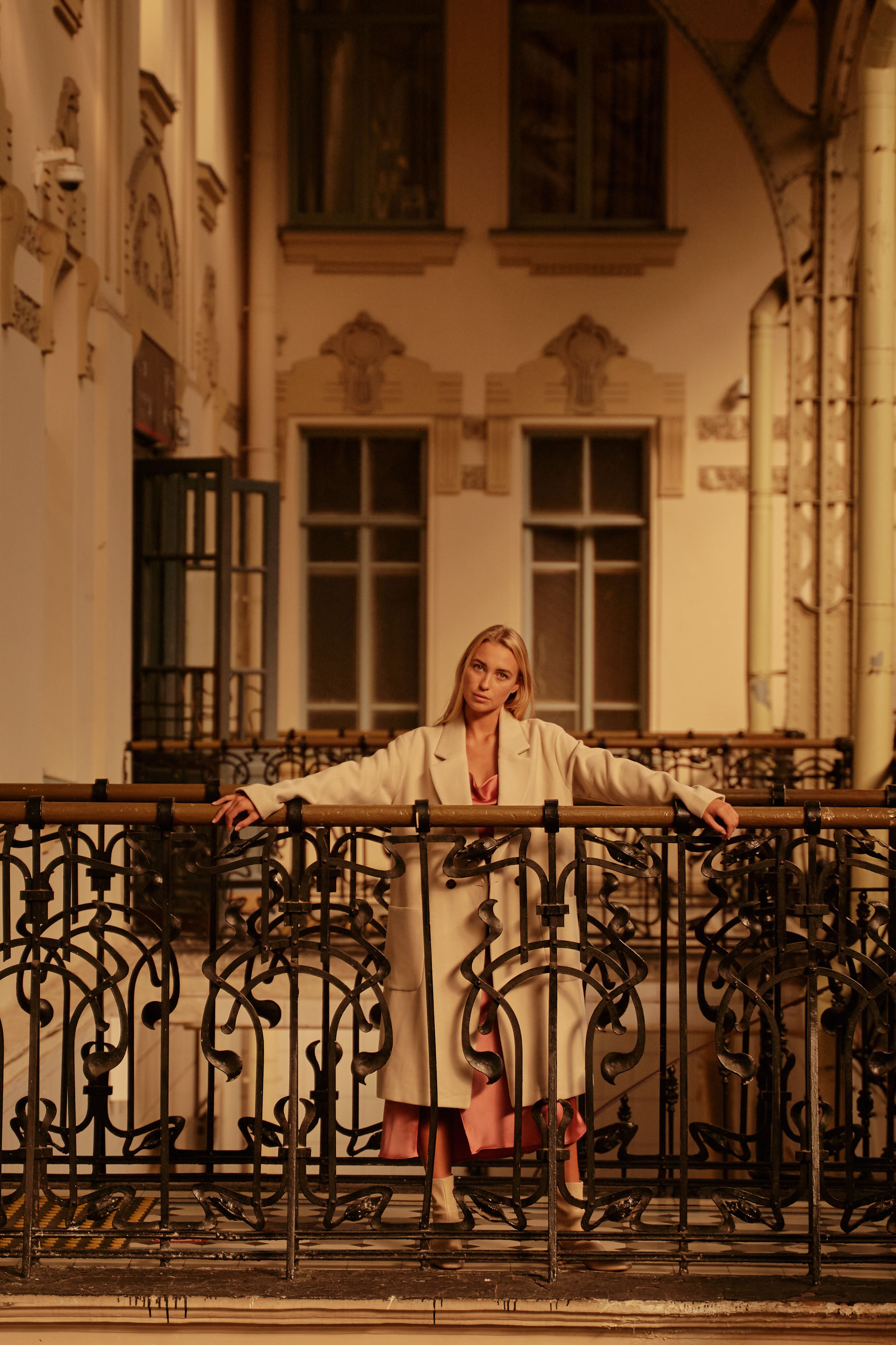 Fashionable woman in elegant white coat posing by ornate metal railing indoors.