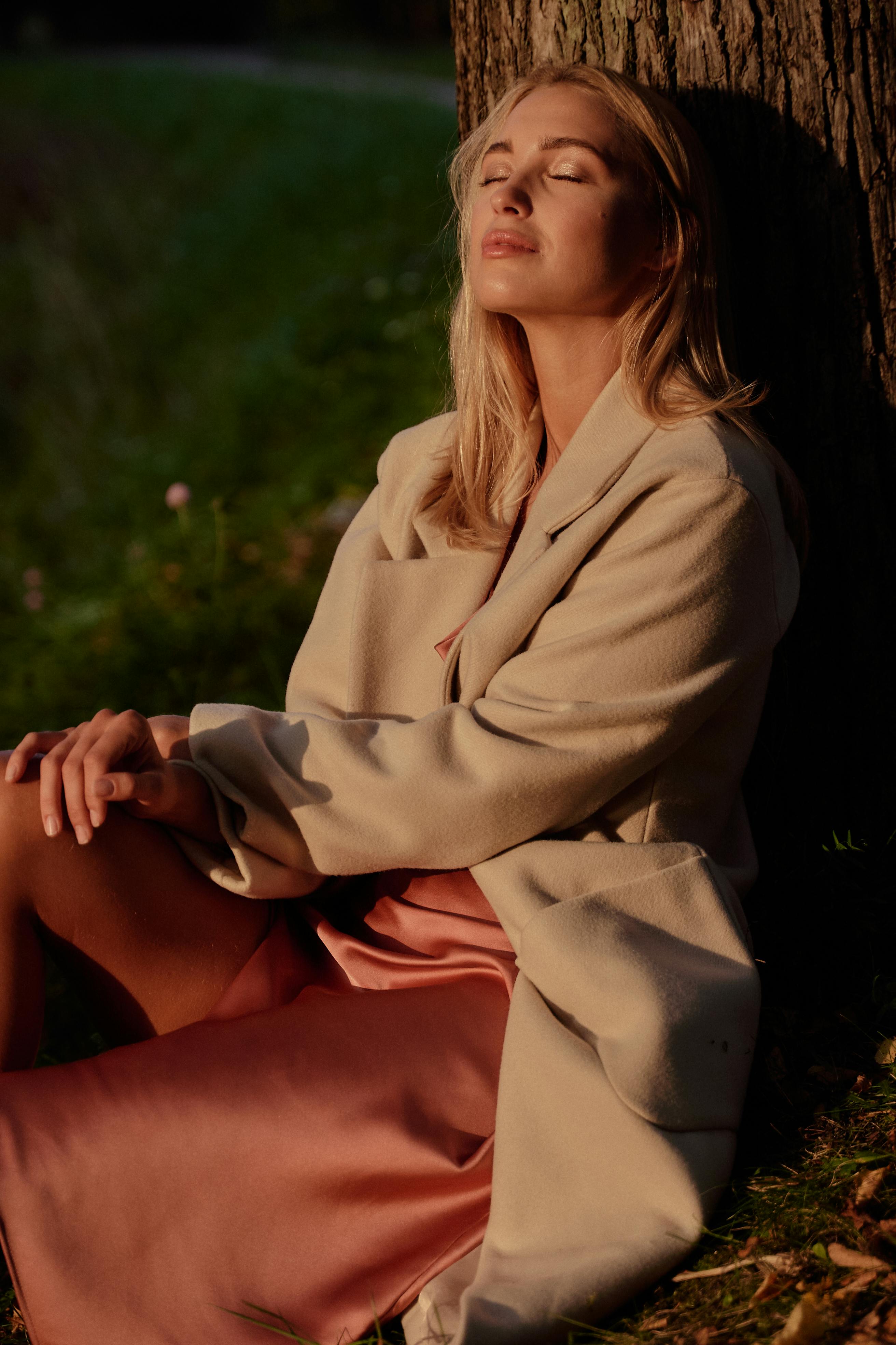 一位金发成年女子穿着米色外套和粉色连衣裙，坐在宁静阳光下的树旁