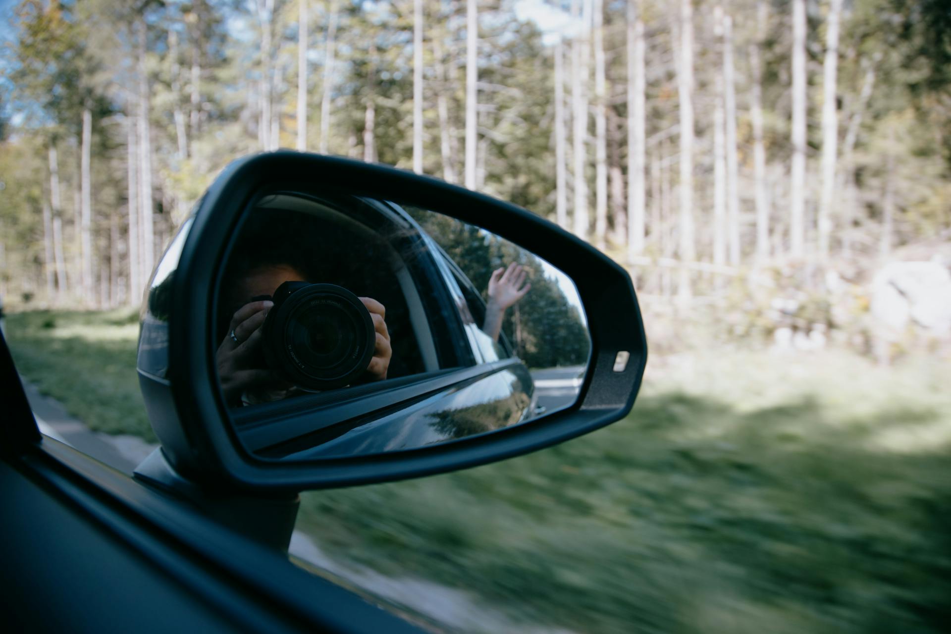 Unrecognizable female holding photo camera and taking selfie through automobile mirror while riding on road between forest