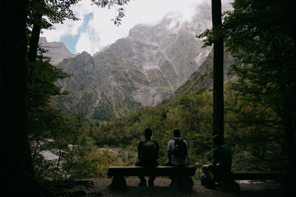 Three hikers seen from behind, sitting together on a bench, resting and taking in a forested mountain view