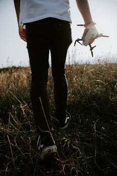 A man walks through a grassy field holding a drone, captured from behind.