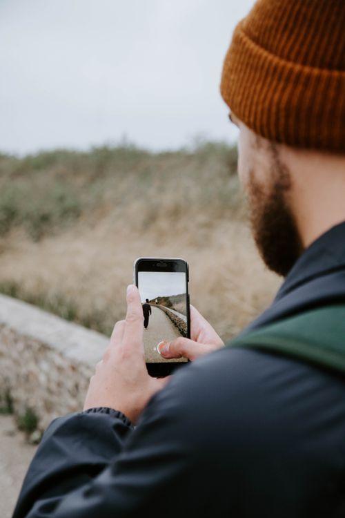Free Back view of bearded man in hat using phone to take photo of friend walking on promenade in countryside Stock Photo