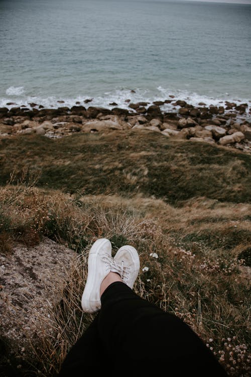 Person in Black Pants and White Sneakers Sitting on Brown Grass Near Body of Water