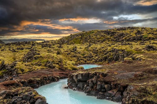 Green and Brown Mountain Beside Body of Water Under Dark Clouds