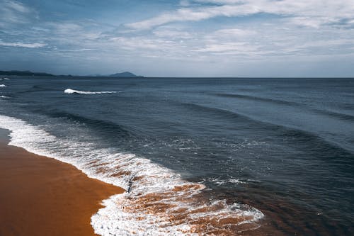 Peaceful scenery of endless ocean with foamy waves rolling on desolated sandy beach on clear day