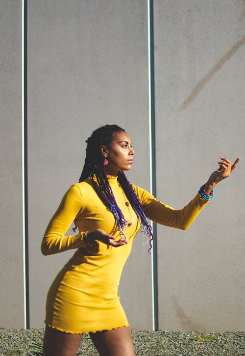 Young black woman in yellow dress and with braids raising hands while standing near building in sunny summer day