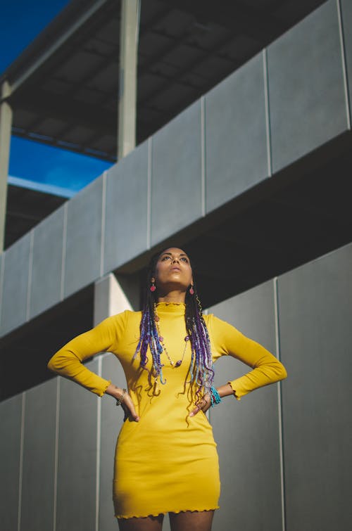 From below of young African American lady standing near construction in yellow dress and with braids and with hand on waist in sunny summer day and looking up